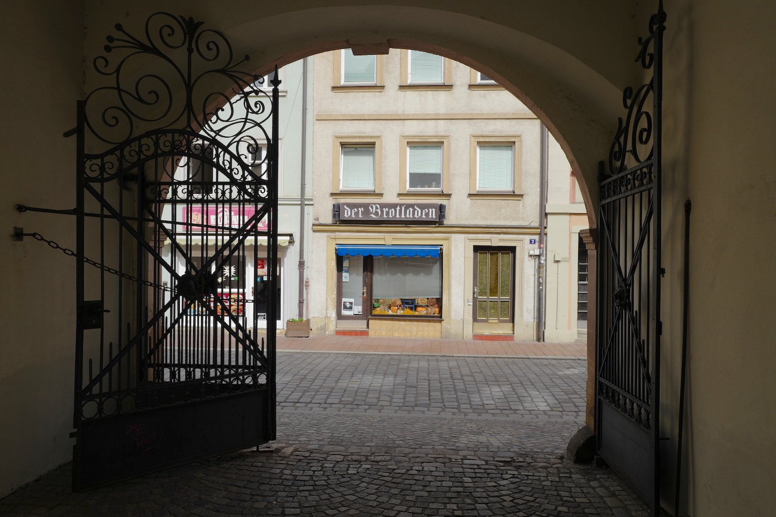 Blick durch ein schmiedeeisernes Tor auf der gegenüberliegenden Straßenseite zum Brotladen